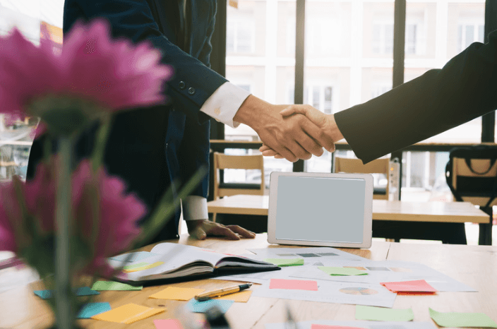 Zwei Geschäftsleute schütteln sich die Hände über einem Schreibtisch mit Tablet, Notizbuch und bunten Klebezetteln.