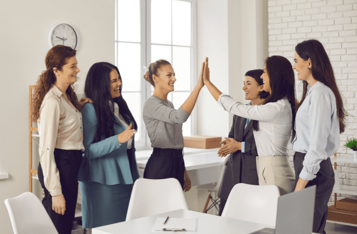 Sechs professionell gekleidete Frauen feiern im hellen Büro einen Erfolg mit einem High-Five.