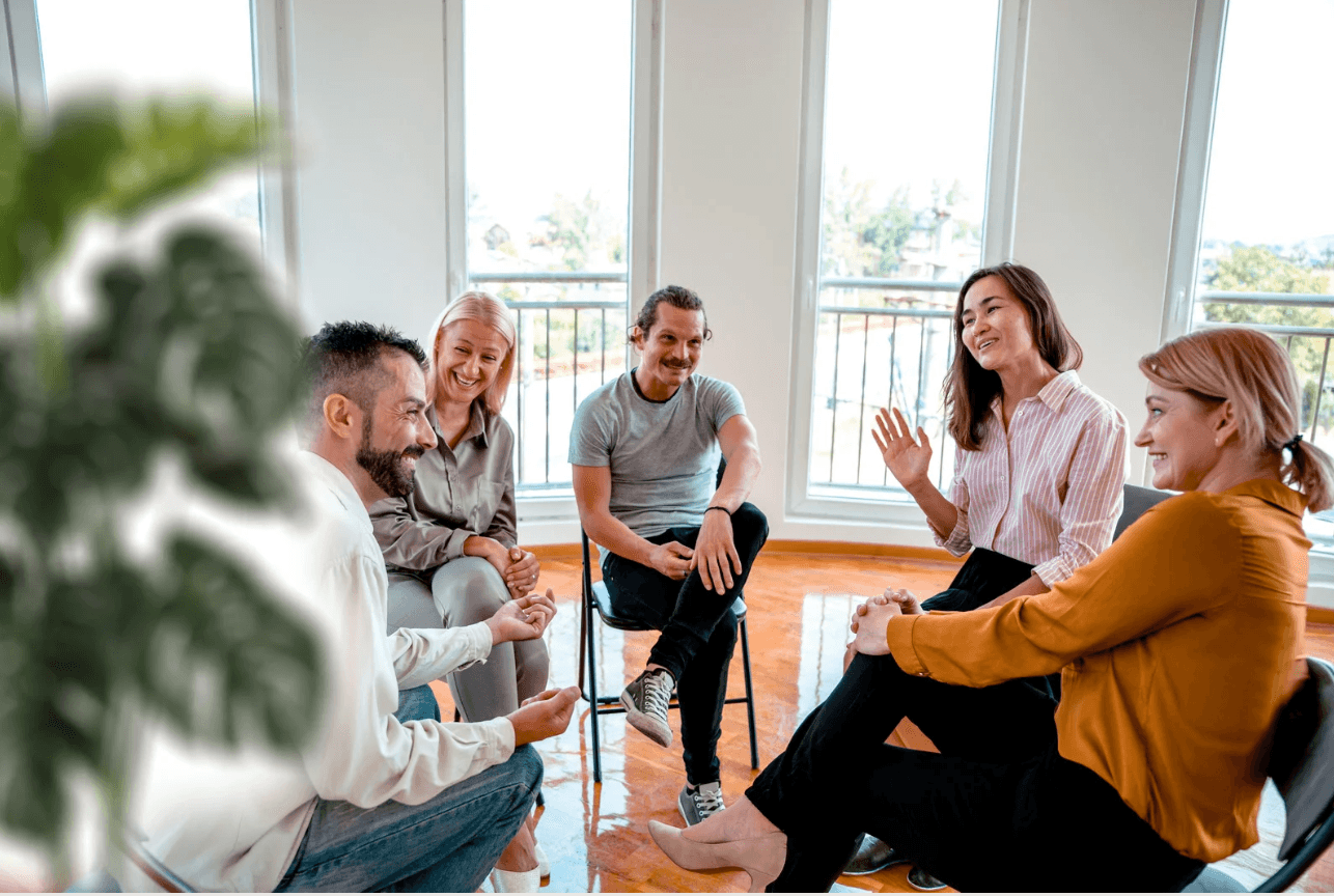 Eine Gruppe von Menschen sitzt im Kreis und unterhält sich lächelnd in einem hellen Raum.