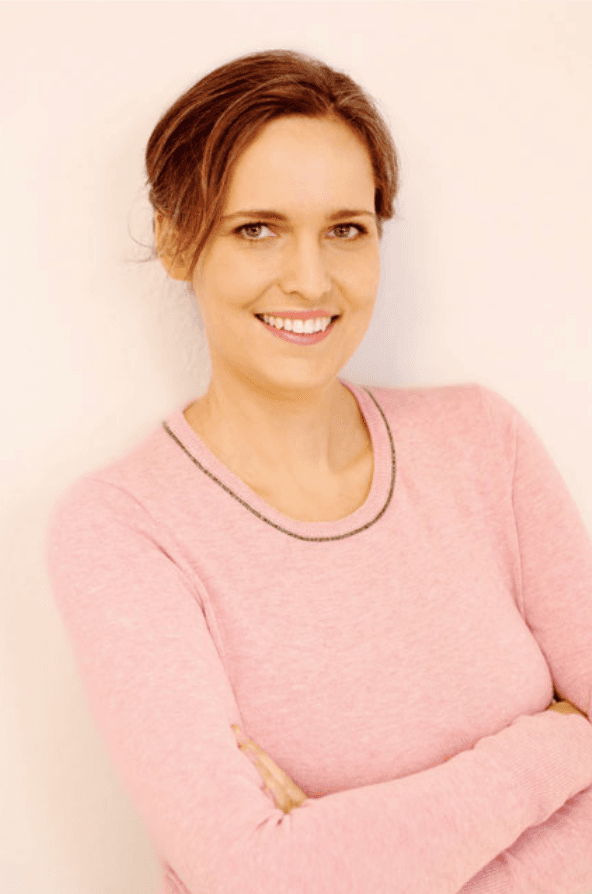 Porträt einer lächelnden Frau mit braunem Haar im rosa Pullover vor weißem Hintergrund.