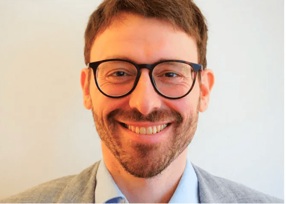 Ein lächelnder Mann mit Brille, kurzem braunem Haar und Bart trägt ein helles Sakko.