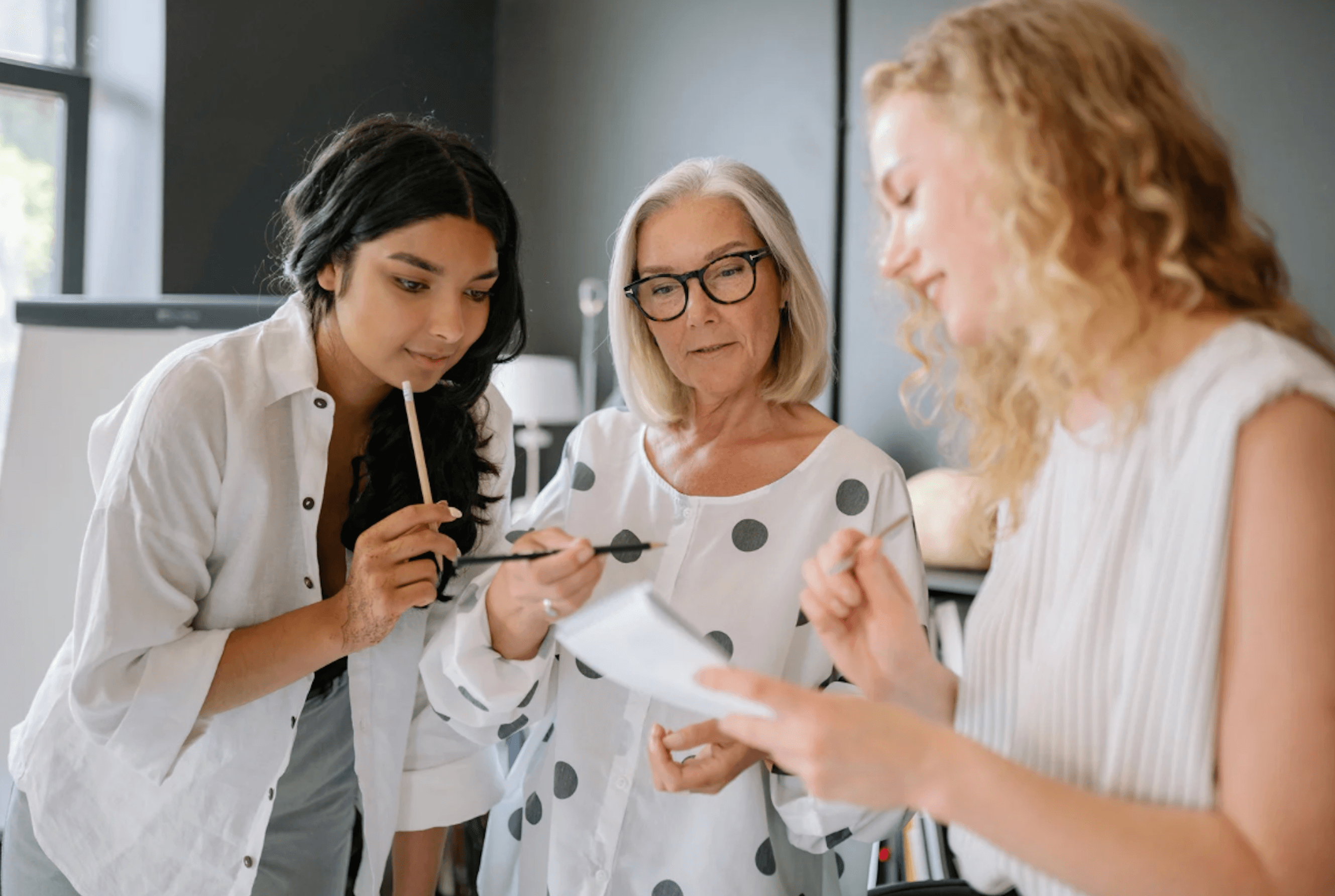 Drei Frauen unterschiedlichen Alters arbeiten gemeinsam an einem Projekt und betrachten konzentriert einen Notizblock.