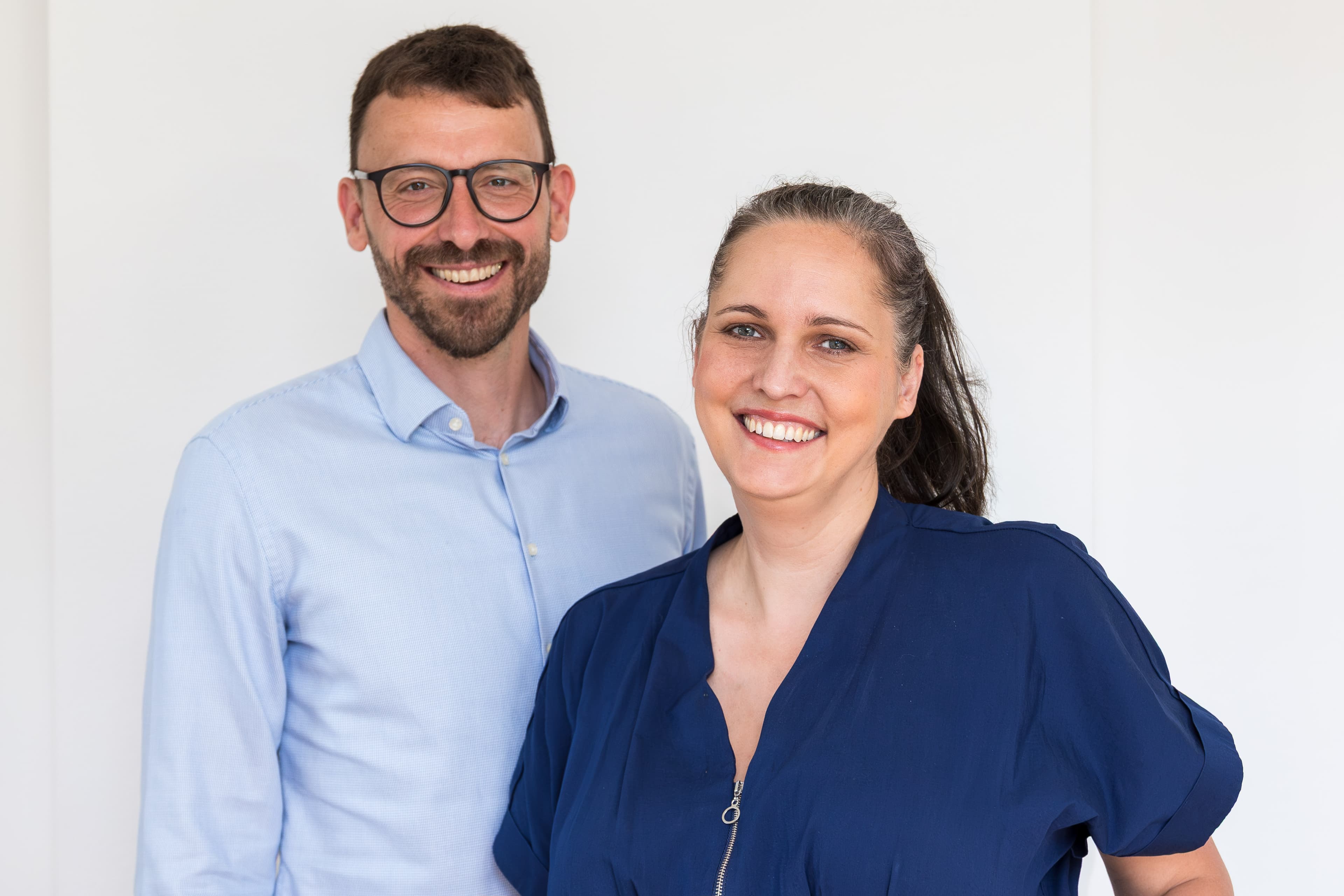 Ein lächelnder Mann mit Brille und eine Frau mit Pferdeschwanz vor einem weißen Hintergrund.